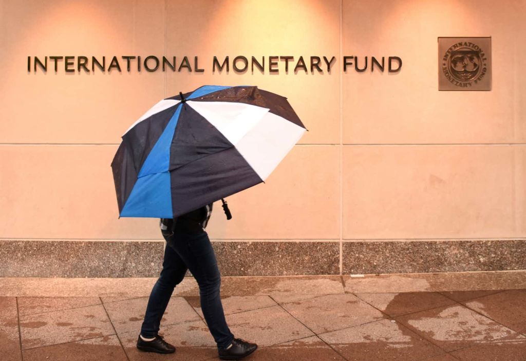 Image of a person walking on the street on a rainy day, covered by a big umbrella with a "International Monetary Fund" sign in the background