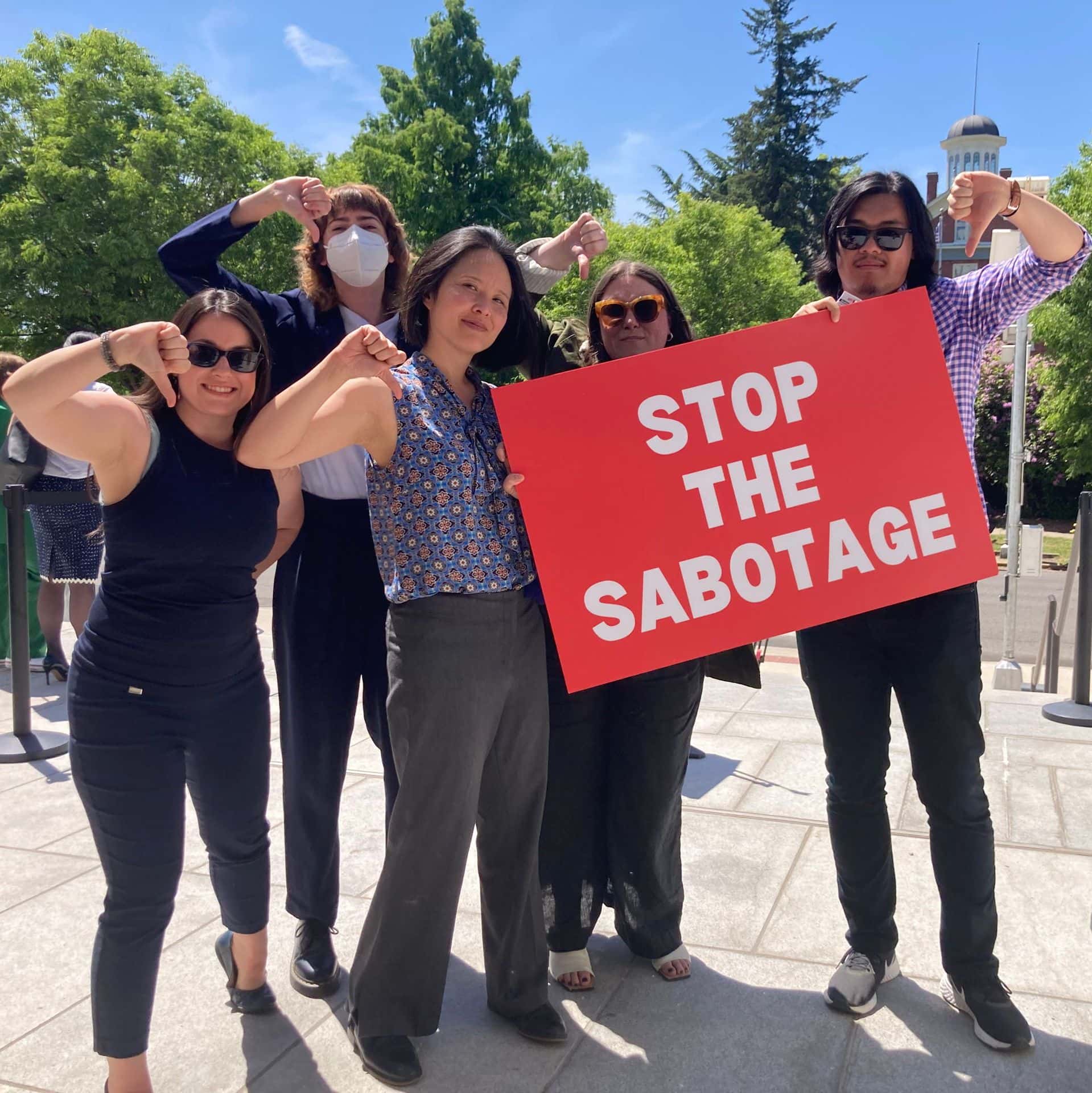 A group of people are posed while holding a sign that reads "Stop the Sabotage." They are all giving the thumbs-down gesture