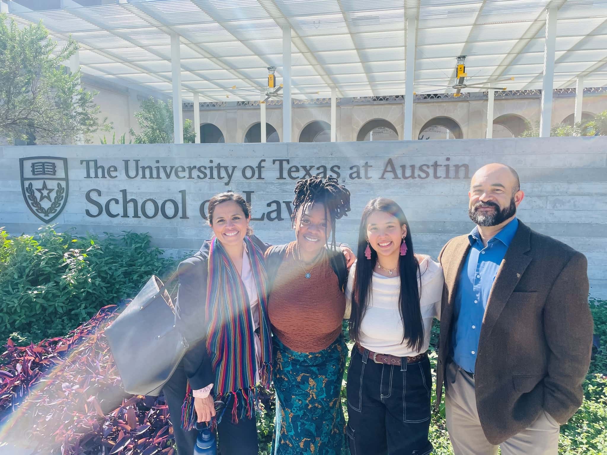 Four people stand in front of a sign that reads "The University of Texas at Austin, School of Law.'