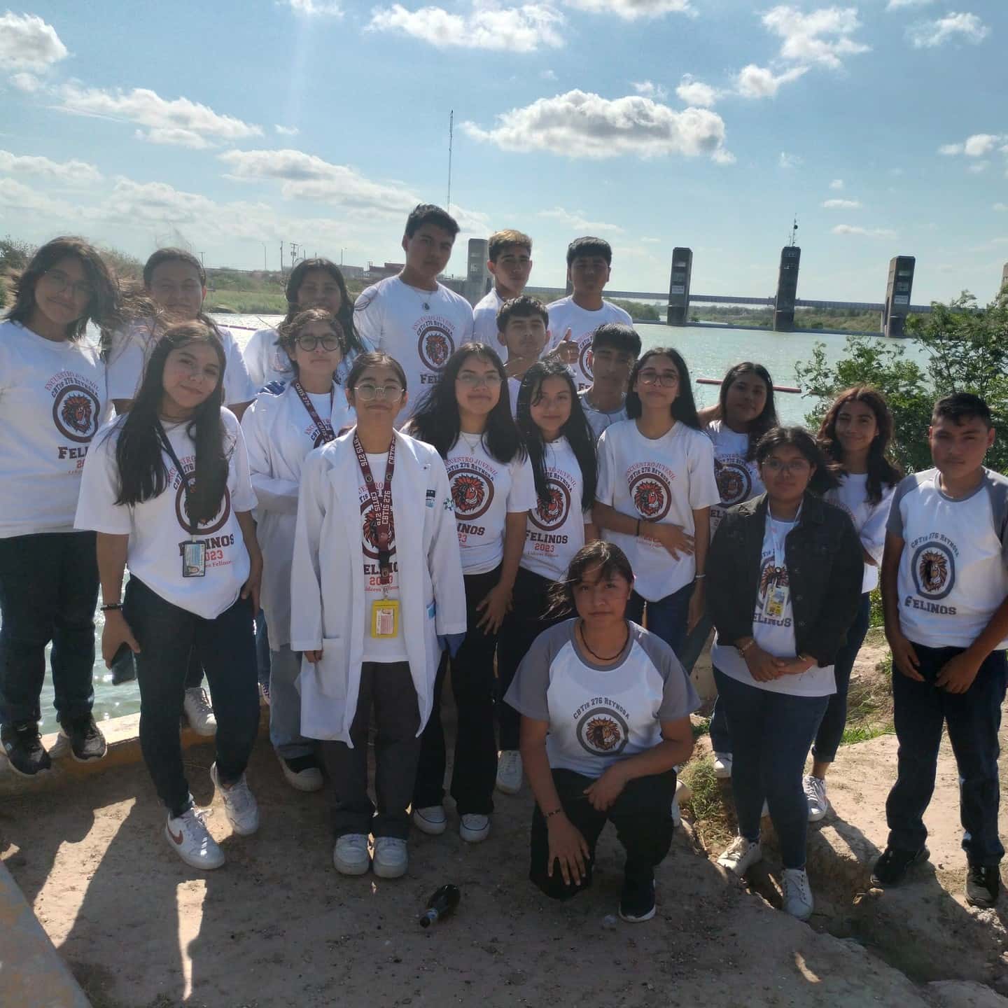A group of students in matching t-shirts stands in front of a waterway for a photo