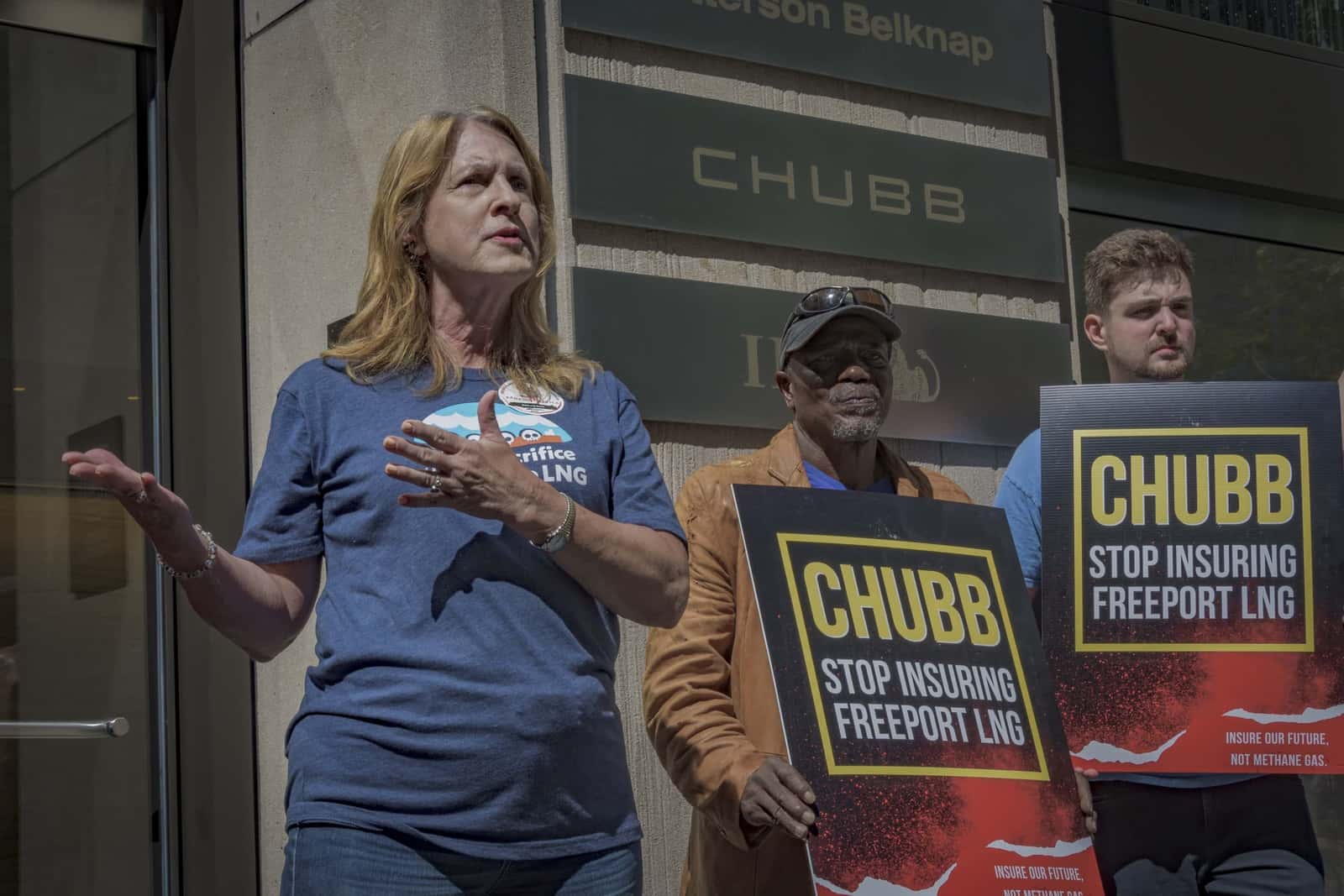 A woman is gesturing with her hands while speaking. Two men standing to her right hold signs that read "CHUBB stop insuring Freeport LNG."