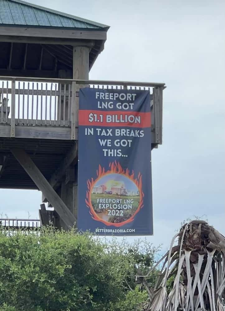 A sign hangs off of the railing of a public beach building that reads, "Freeport LNG got .1 Billion in tax breaks, we got this.." and depicts a photo of the Freeport LNG facility exploding