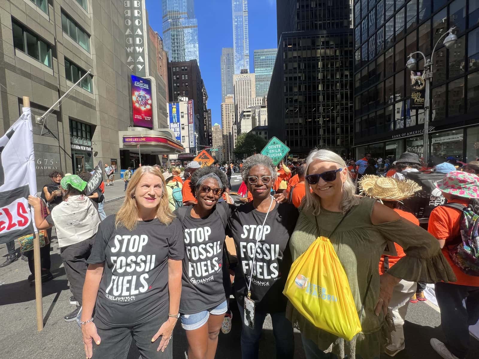 Four women stand on a city sidewalk in bright sunlight, wearing matching black t-shirts that read "Stop Fossil Fuels."
