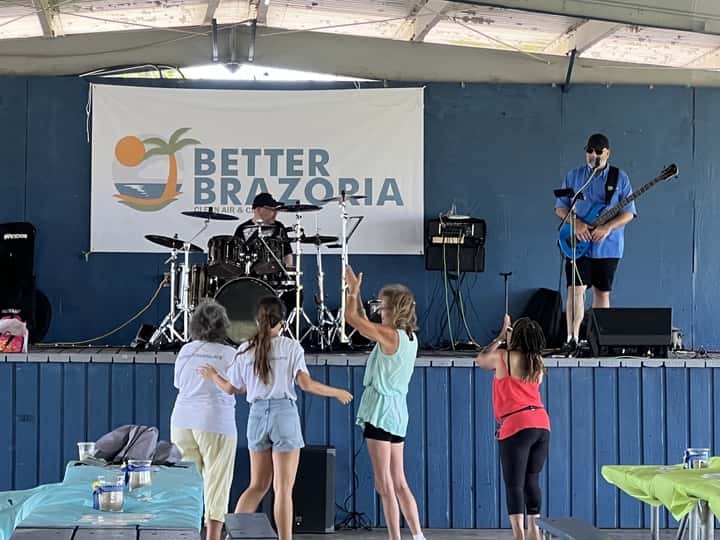 A small group of people are gathered in front of a blue stage, where a drummer and bassist are playing in front of a sign that reads "Better Brazoria"