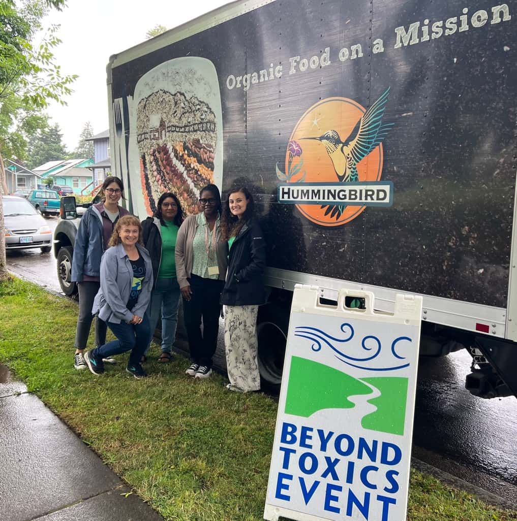 A group of people stand in front of a truck with graphics that read "Organic Food on a Mission." A sign on the ground reads "Beyond Toxics Event"