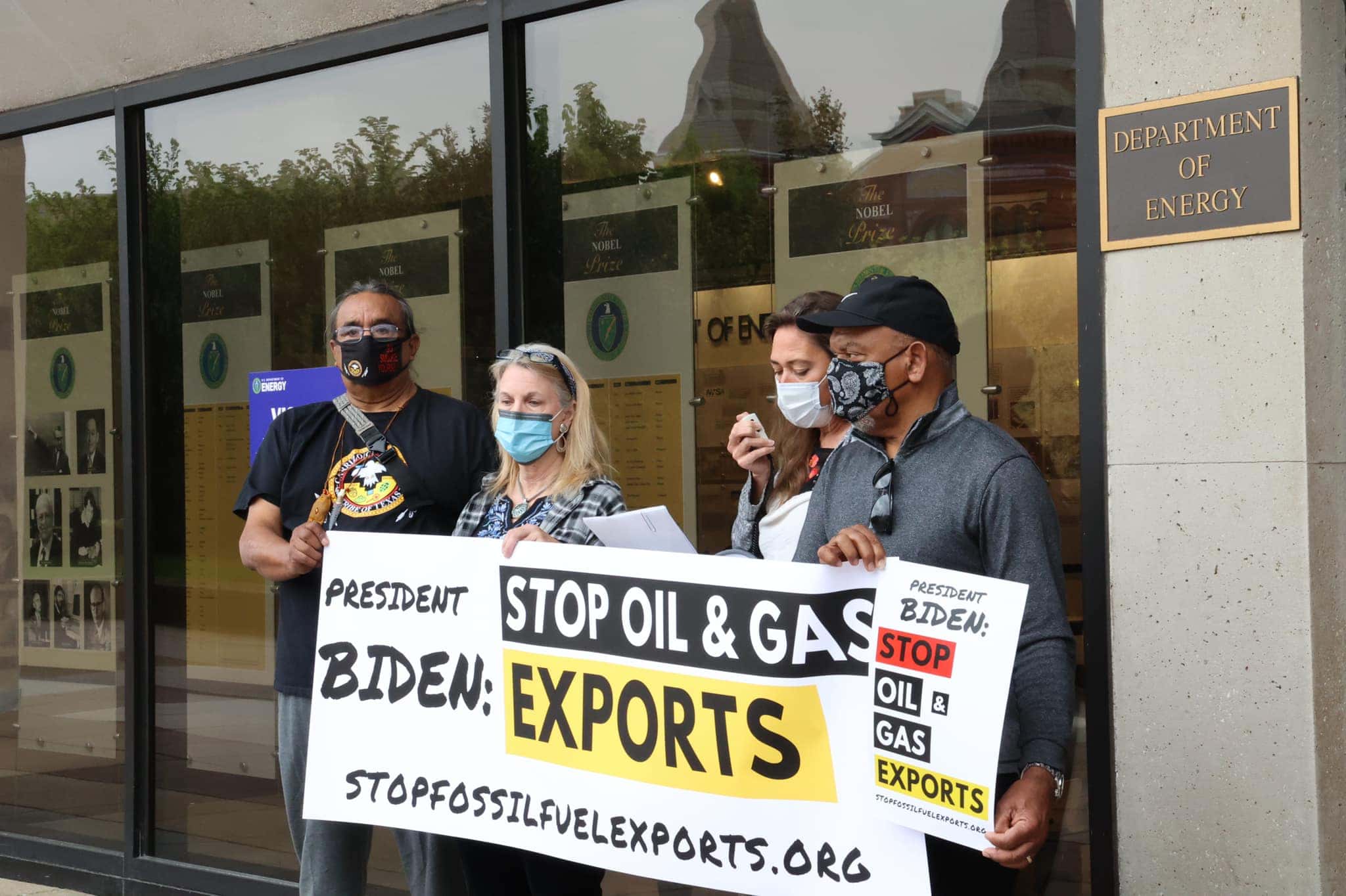 A group of four people stand holding a protest banner that reads, "President Biden, stop oil & gas exports."