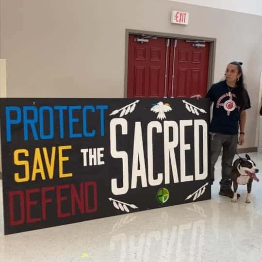A person stands near a hand-made sign that reads "Protect Save Defend the Sacred."