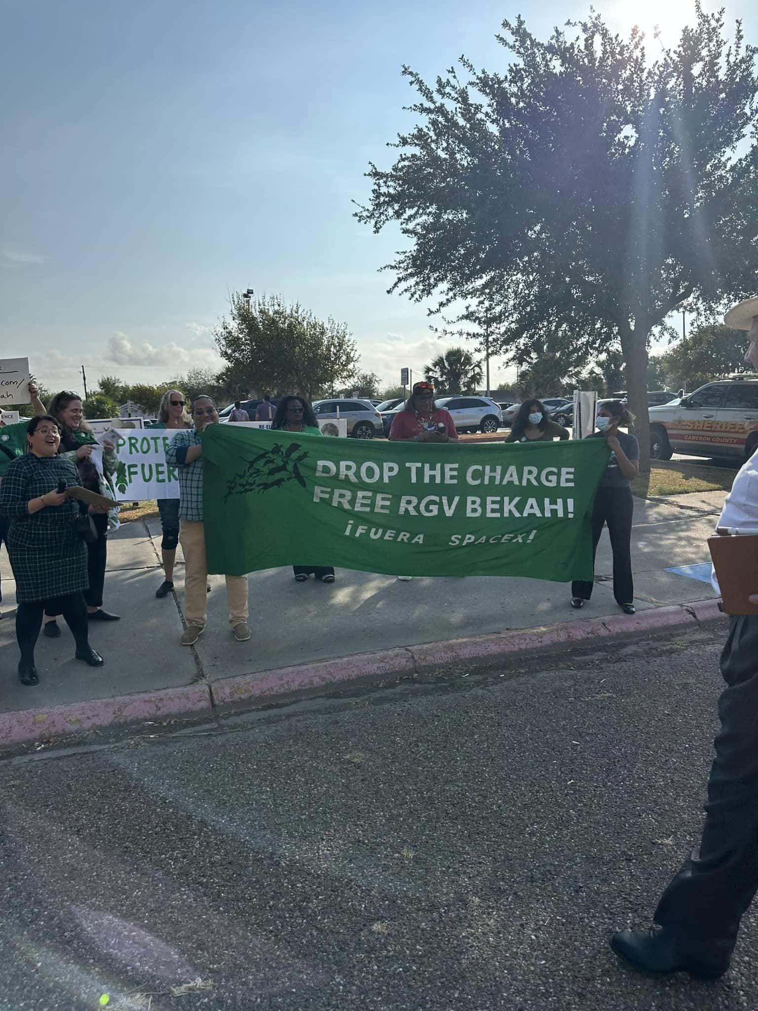 A group of protesters stands together on a street, holding a green banner that reads "DROP THE CHARGE FREE RGV BEKAH!"