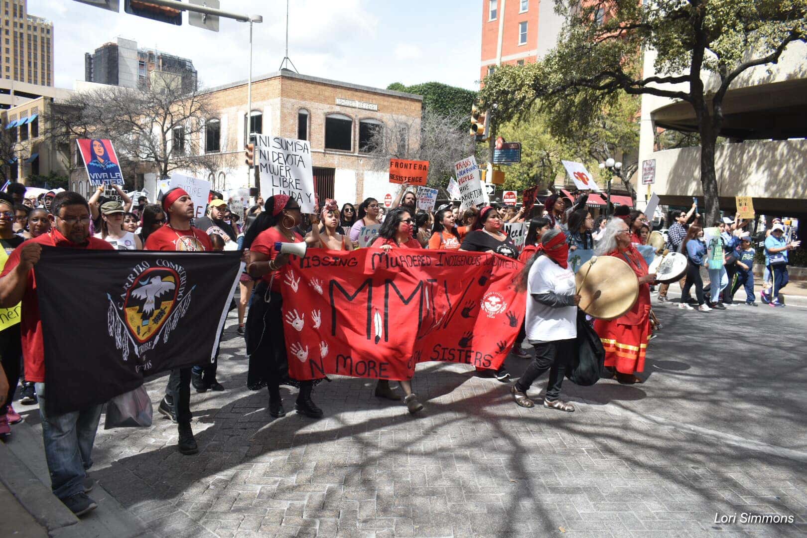 A group of Carrizo Comecrudo Tribe Members marches down the street while holding a hand-made sign.