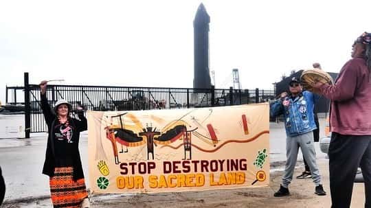A woman and a man are holding a hand-made banner that reads "Stop Destroying our Sacred Lands" in front of a waterway.