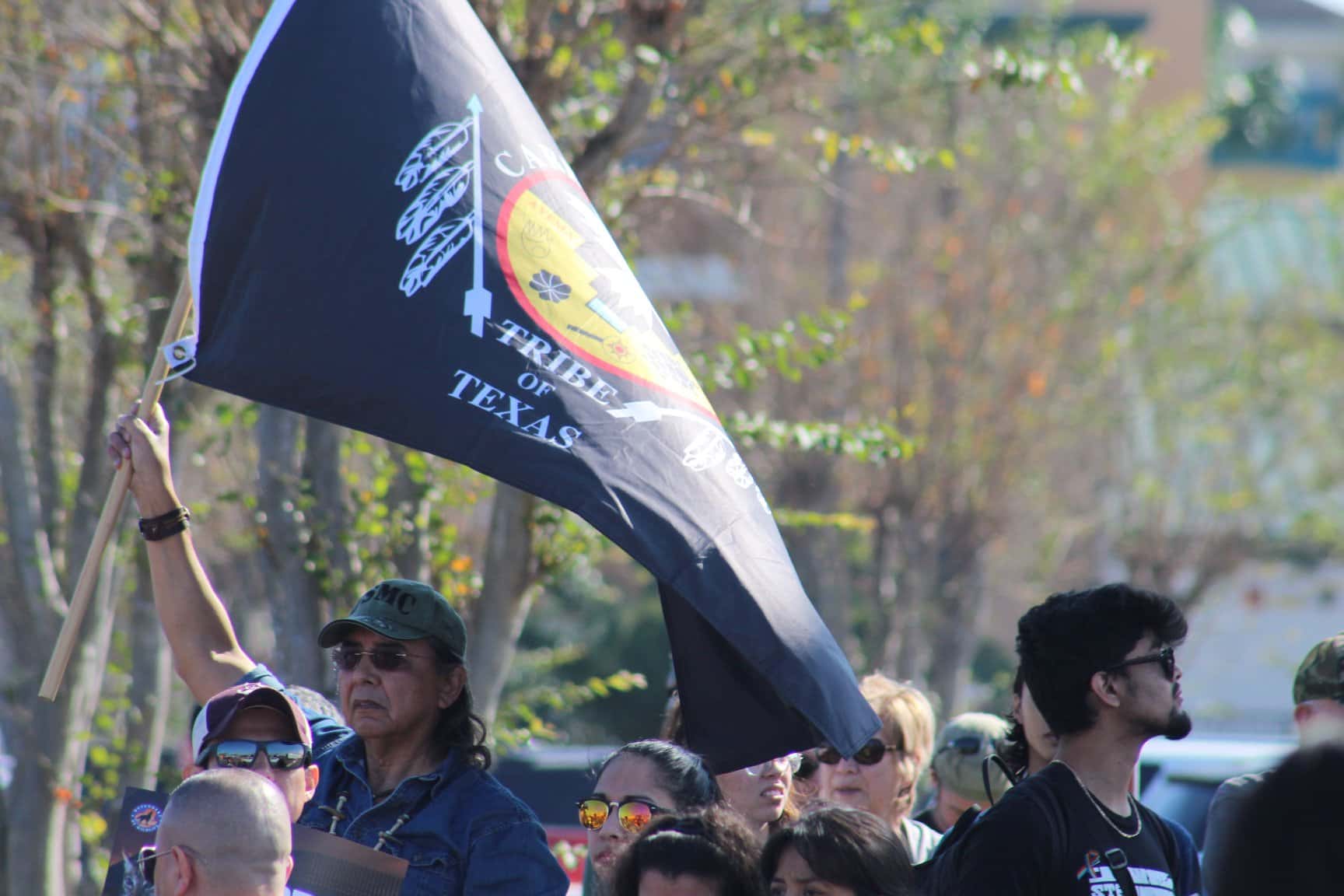 A group of people are gathered in protest. A man waves a black flag with the Carrizo Comecrudo logo above his head. 