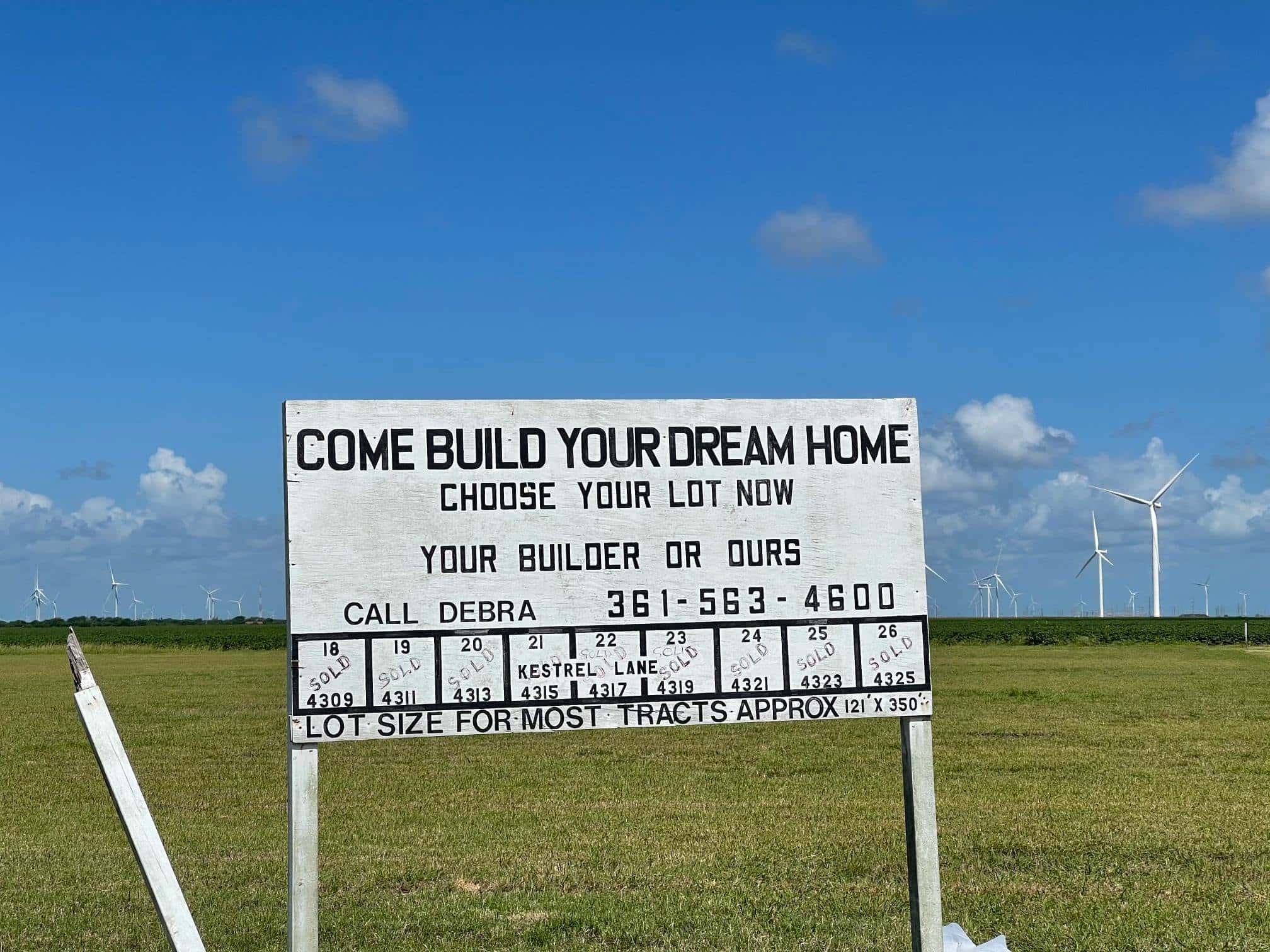 A lone sign stands in an empty field that reads, "Come build your dream home, choose your lot now. Your builder or ours."