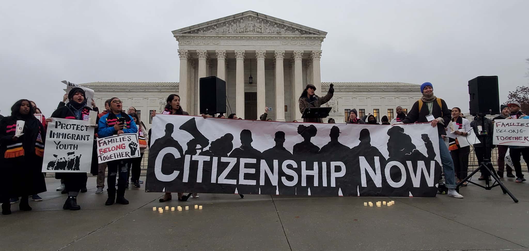 A group of people standing in front of a government building, holding a sign that reads "Citizenship Now."