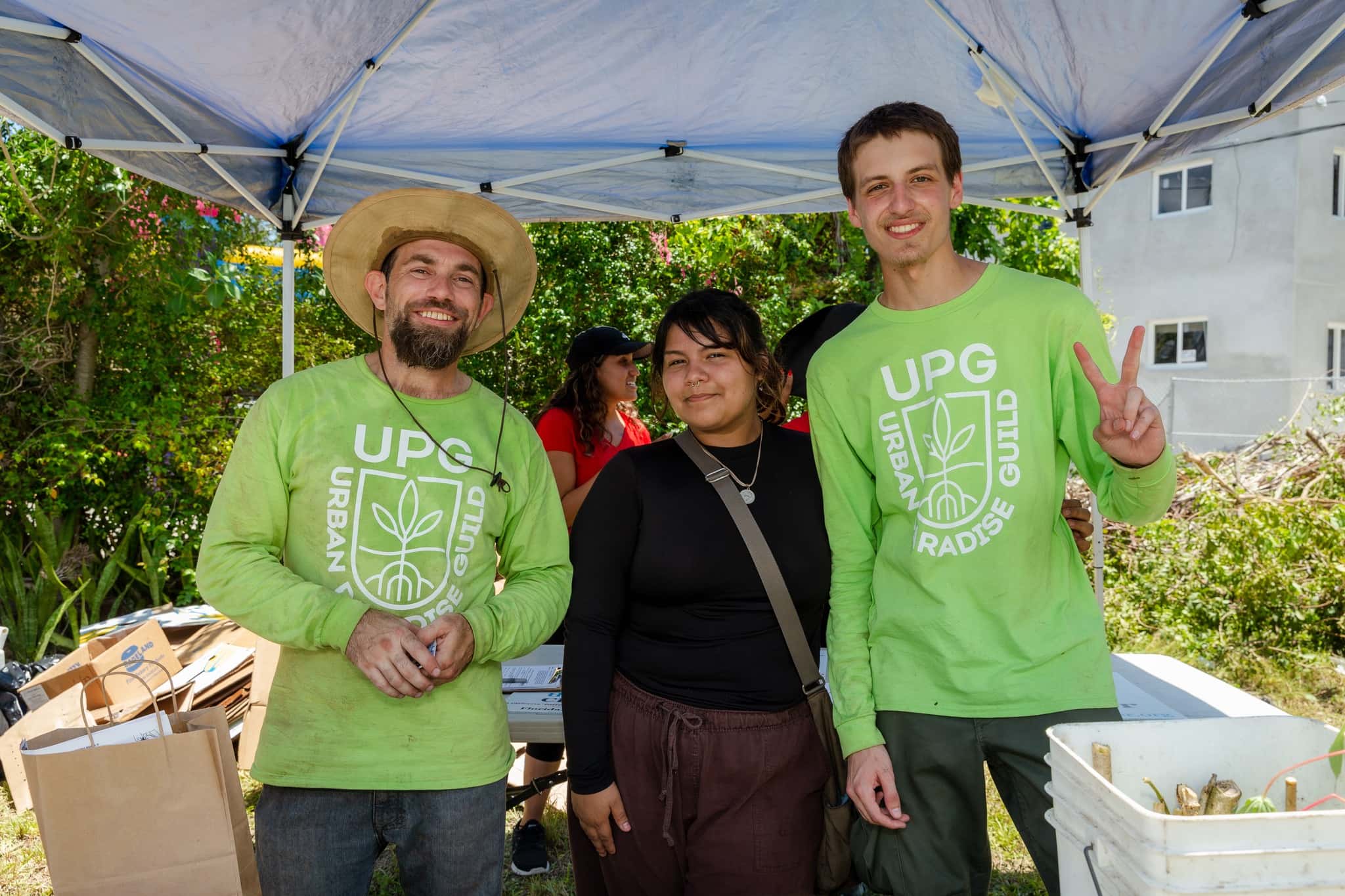 Two men in green shirts stand with a woman in a black shirt under a temporary awning