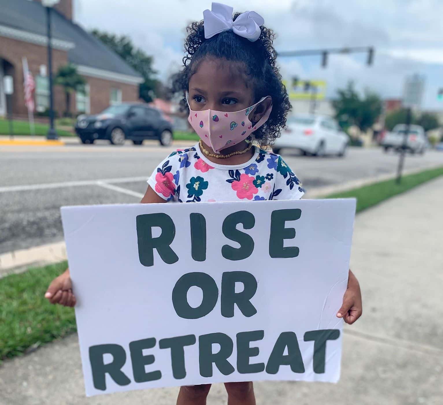 A small girl with a purple bow in her hair holds a sign that reads "Rise or Retreat"