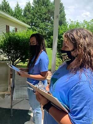 Two women wearing protective masks standing on a porch and holding clipboards