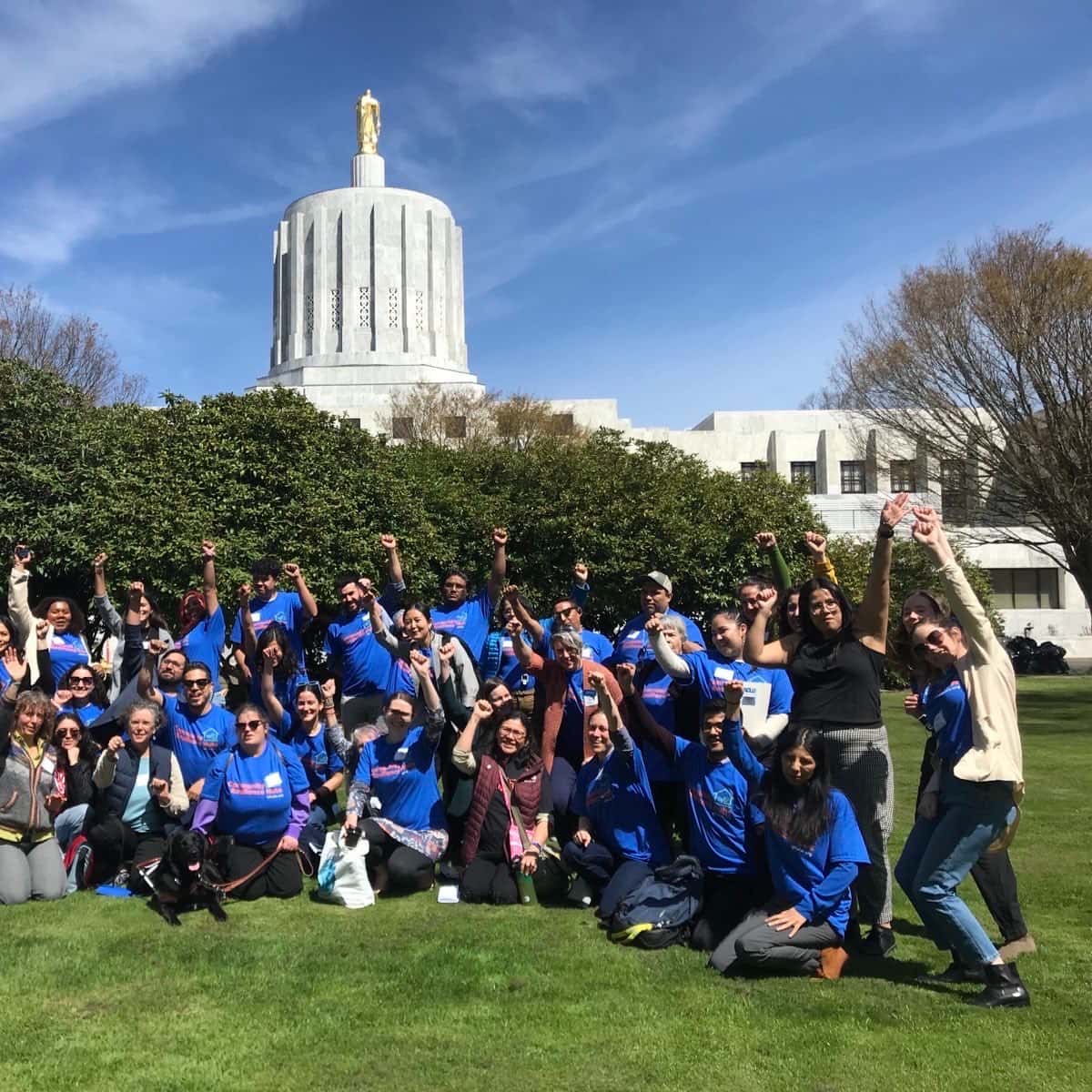 A large group of people in matching blue t-shirts pose on the lawn of a government building for a photo.