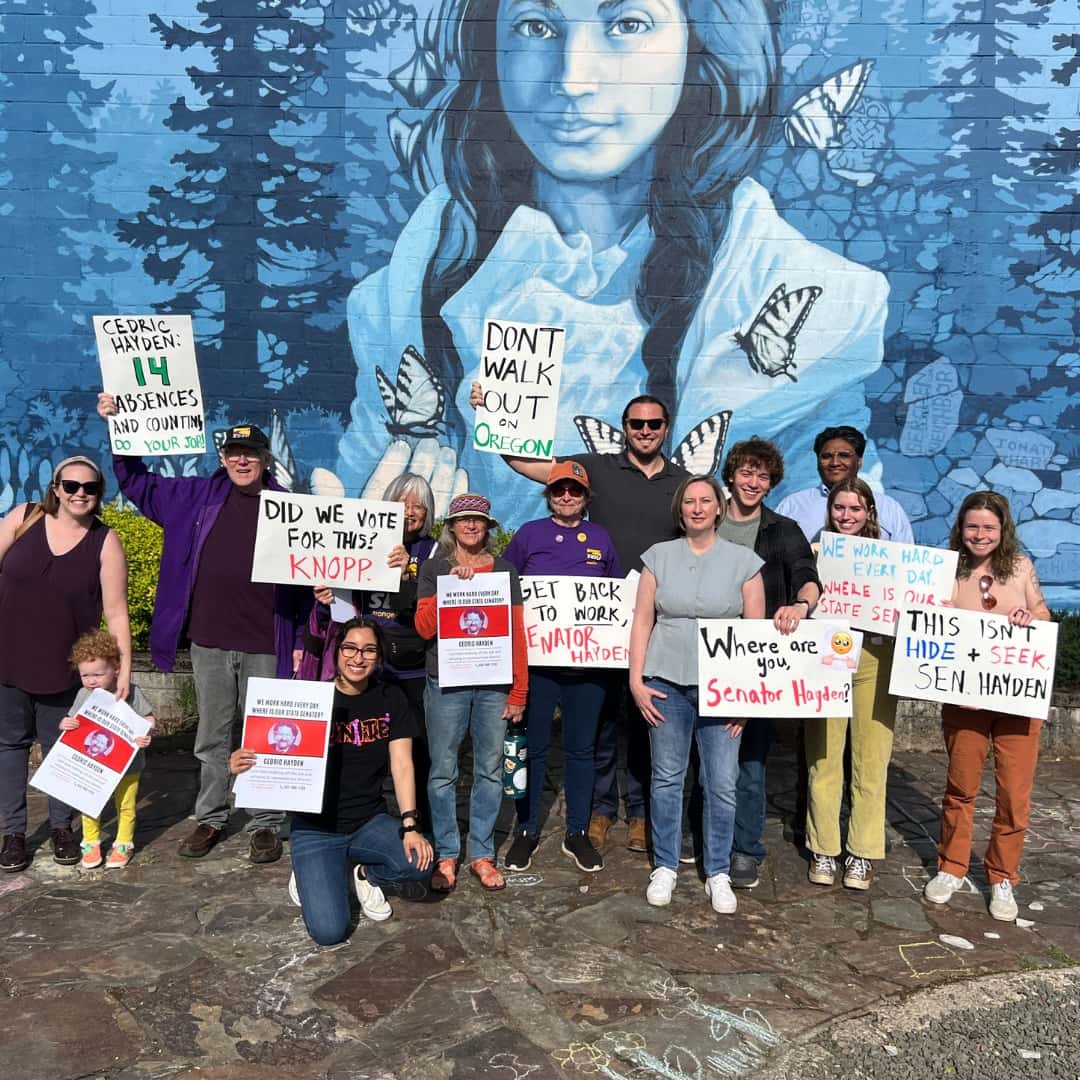 A group of young people stand in front of a mural of an Indigenous woman in a forest surrounded by 2 monarch butterflies. 