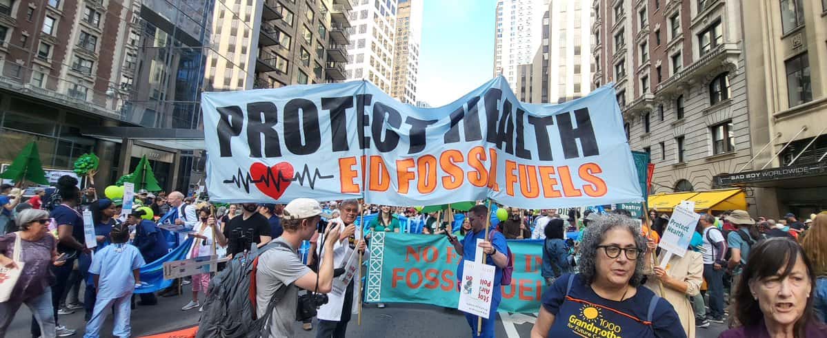 A group of marching protesters in the streets on New York City holding a sign that reads "Protect Health, End Fossil Fuels."