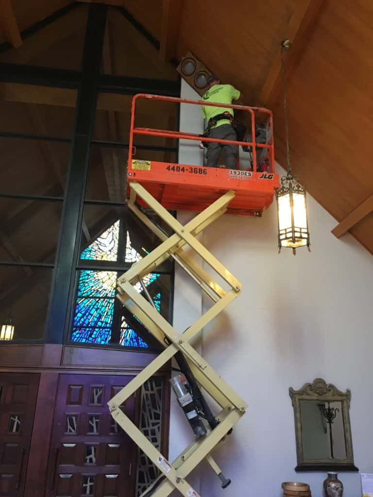 A man on a scissor-lift changes the light fixture attached to the roof of a church.