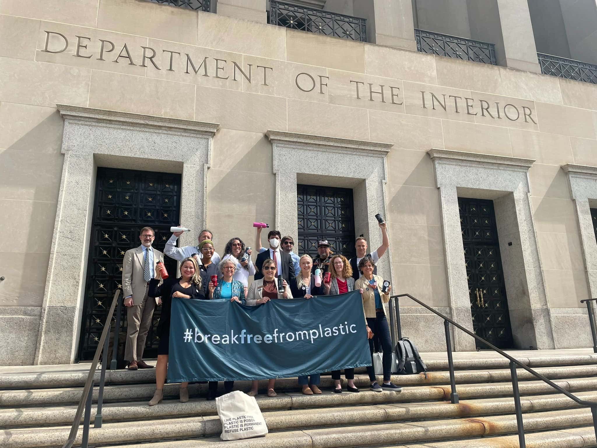 A group of people stands on the steps of the Department of the Interior, holding a black banner that reads "#breakfreefromplastic"