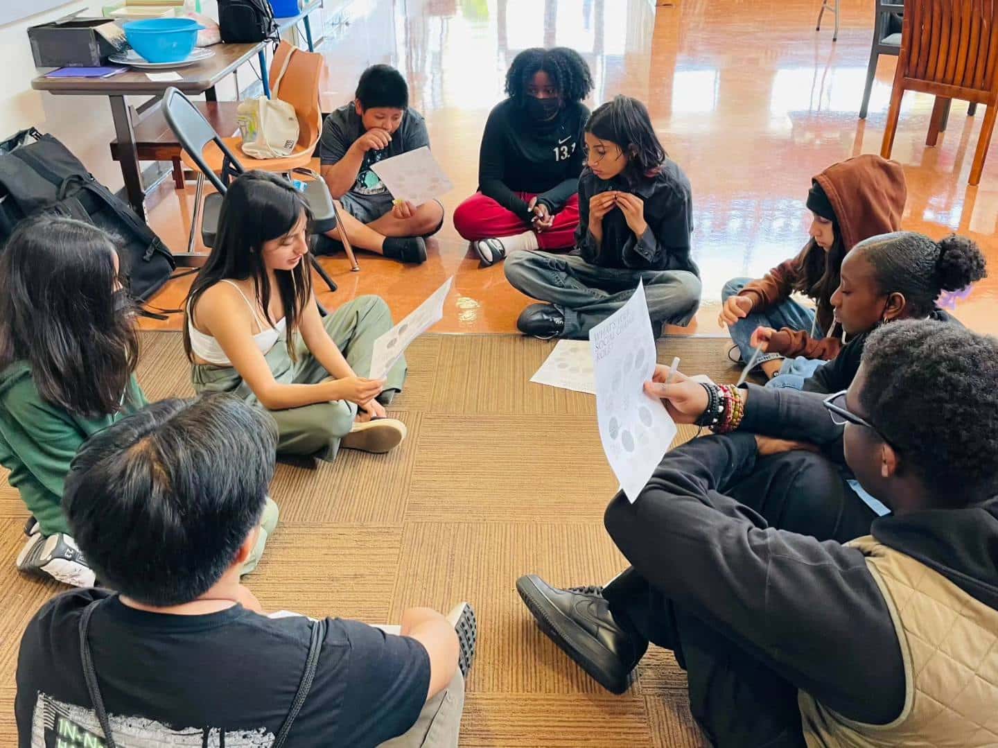 A group of students are sitting on the floor of a classroom, while one student reads a document to them aloud.